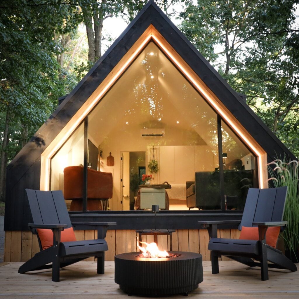 The Marburg Cabin at dusk, its A-frame gable window glowing warm, with a lit fire pit and chairs on the deck